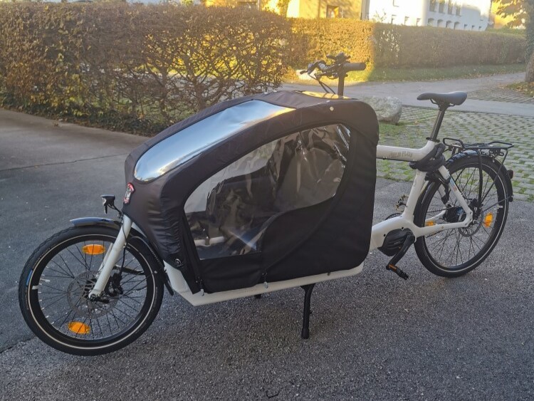 Weißes Lastenrad Emma mit schwarzer Transportbox steht auf einem Asphaltweg vor einer Hecke